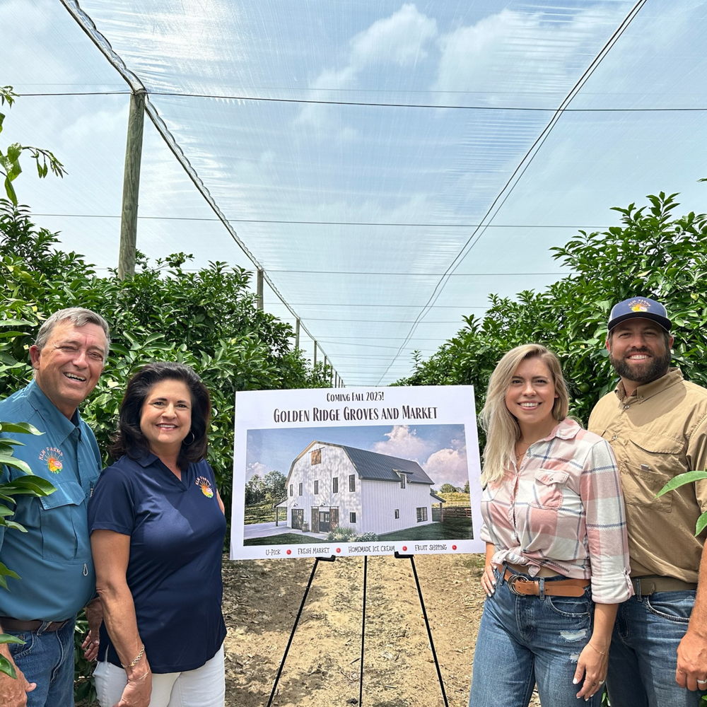 Four people standing outdoors with a sign showing plans for Golden Ridge Groves and Market.